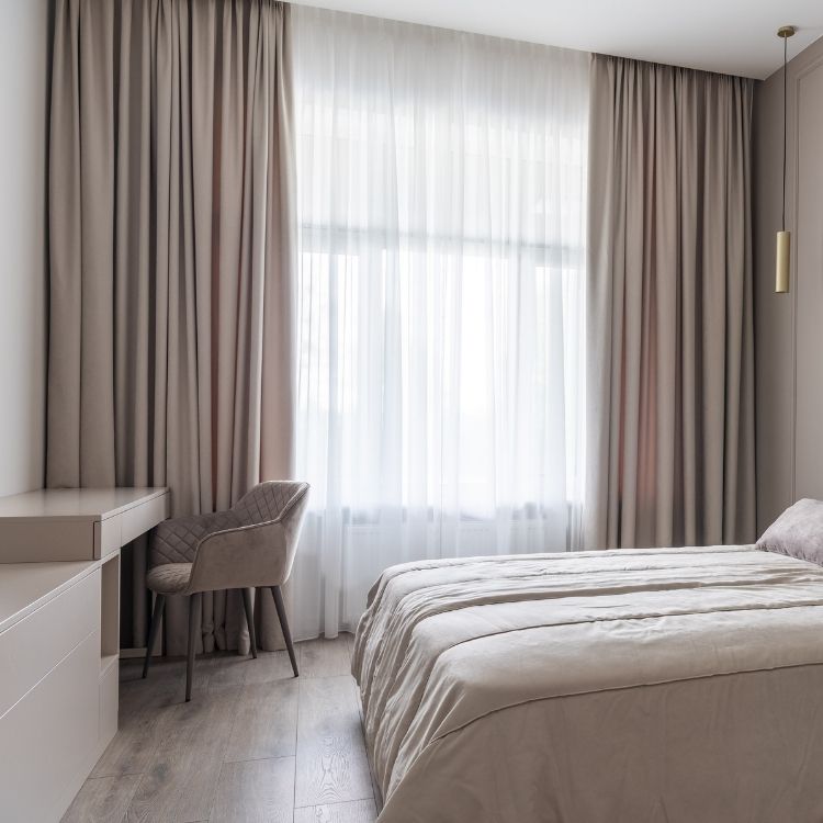 Modern bedroom with soft beige curtains, a cozy bed, and a minimalist desk with a plush chair, bathed in natural light.