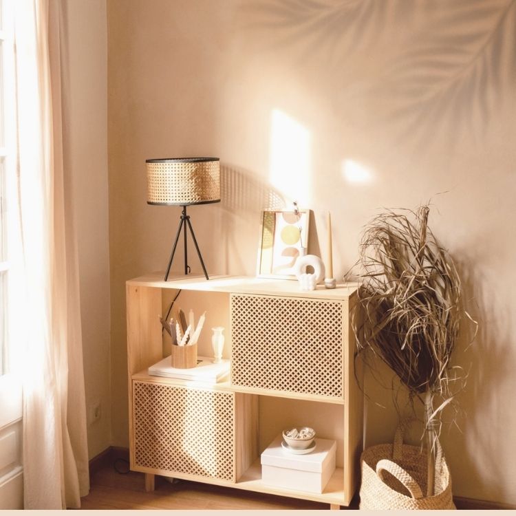 A cozy corner featuring a wooden shelf with decor items, a patterned lamp, and a dried plant, illuminated by soft sunlight.